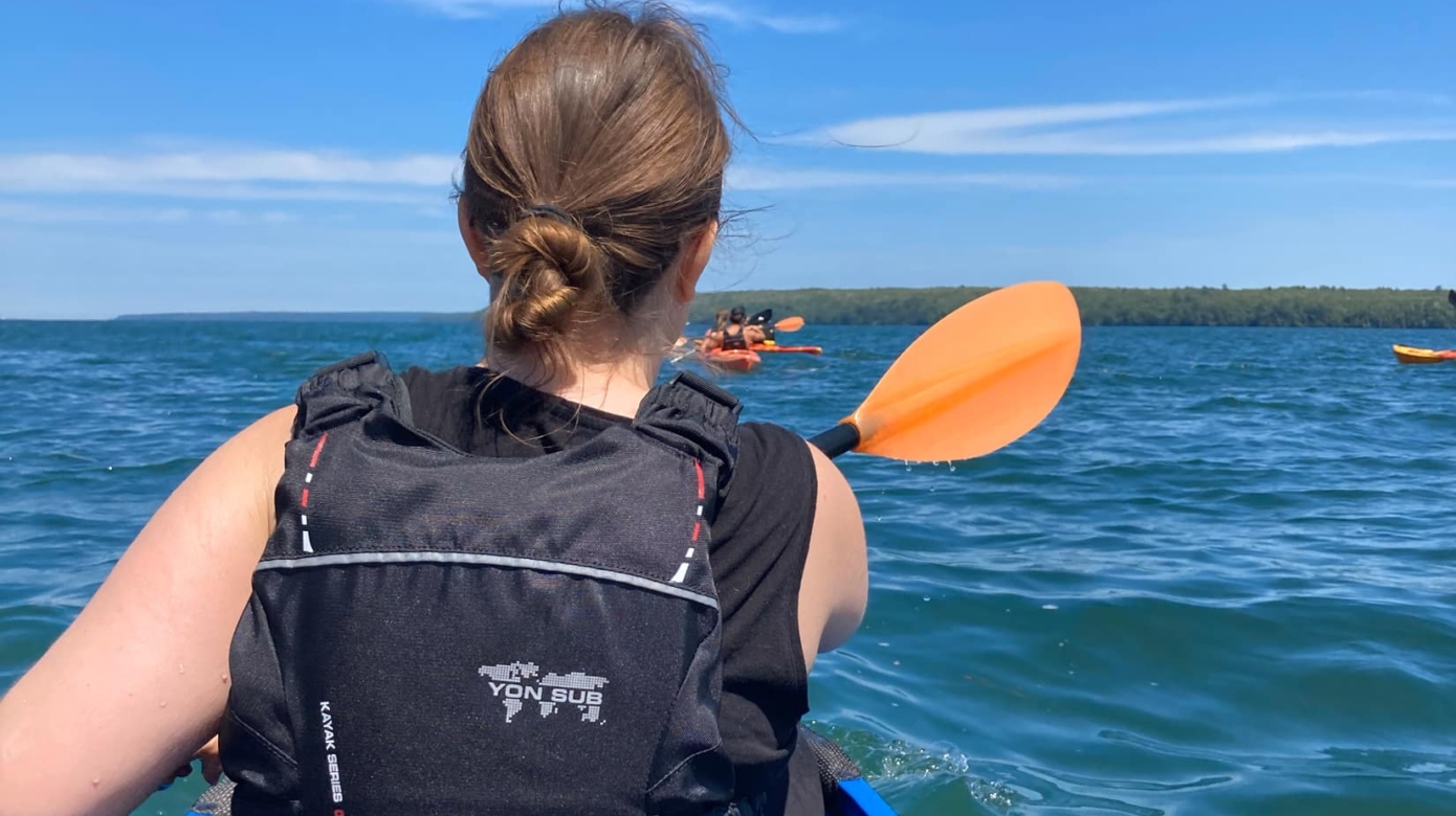 Mary Belle Zook pictured from behind, paddling a kayak on Lake Superior. Her hair is in a bun at the nape of her neck and she wears a black life vest. Blue water meets blue sky beyond her, and small orange kayaks are visible further out on the lake.