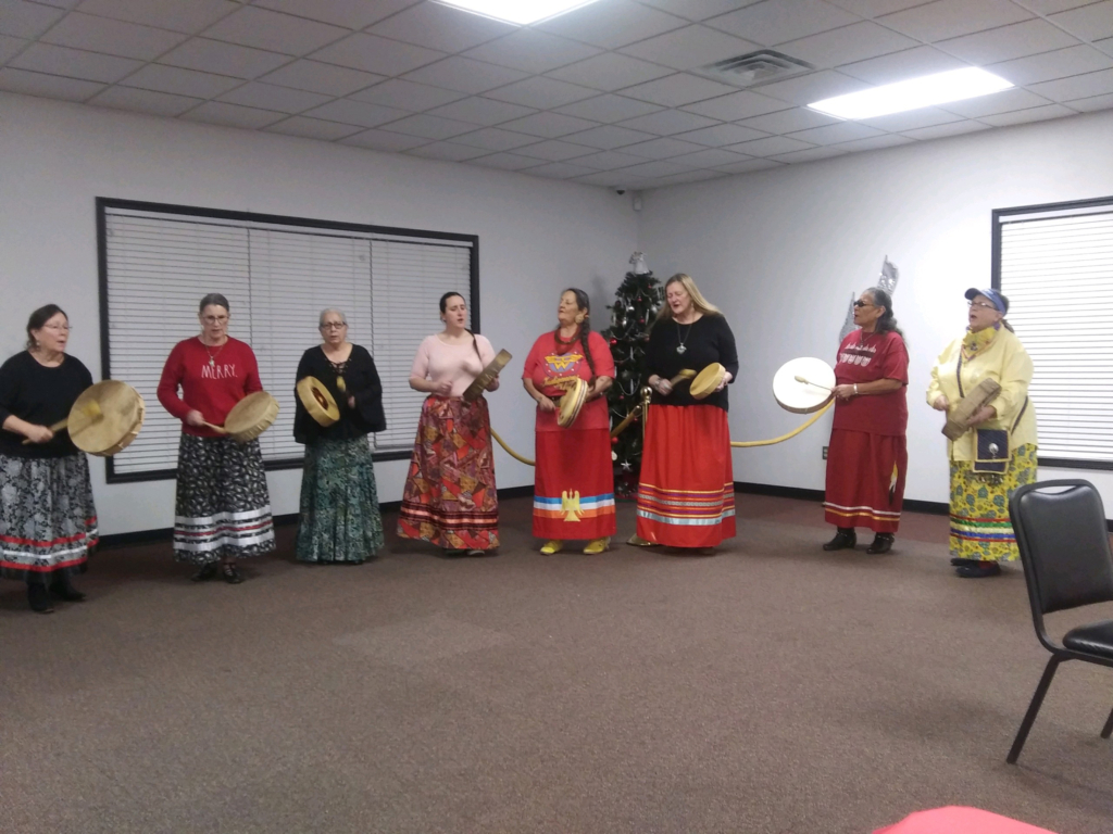 Photo of eight women with hand drums and wearing ribbon skirts in red and green hues at the CPN Veterans Organization Christmas Party 2022.