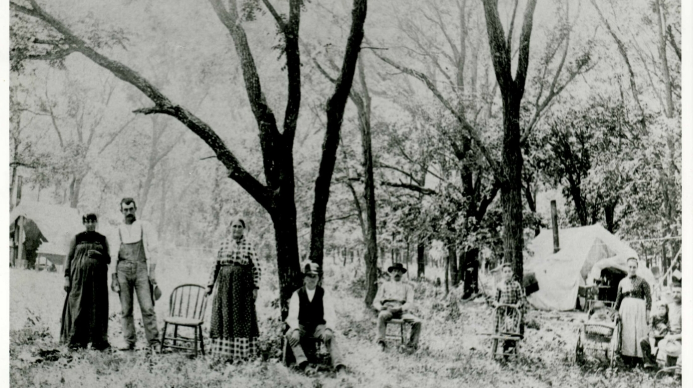 An archival photo of Lousia Smith Storm Hartman and her family from 1898 or 1899. The family is spread out below several trees, leaning on chairs and standing in small groups.