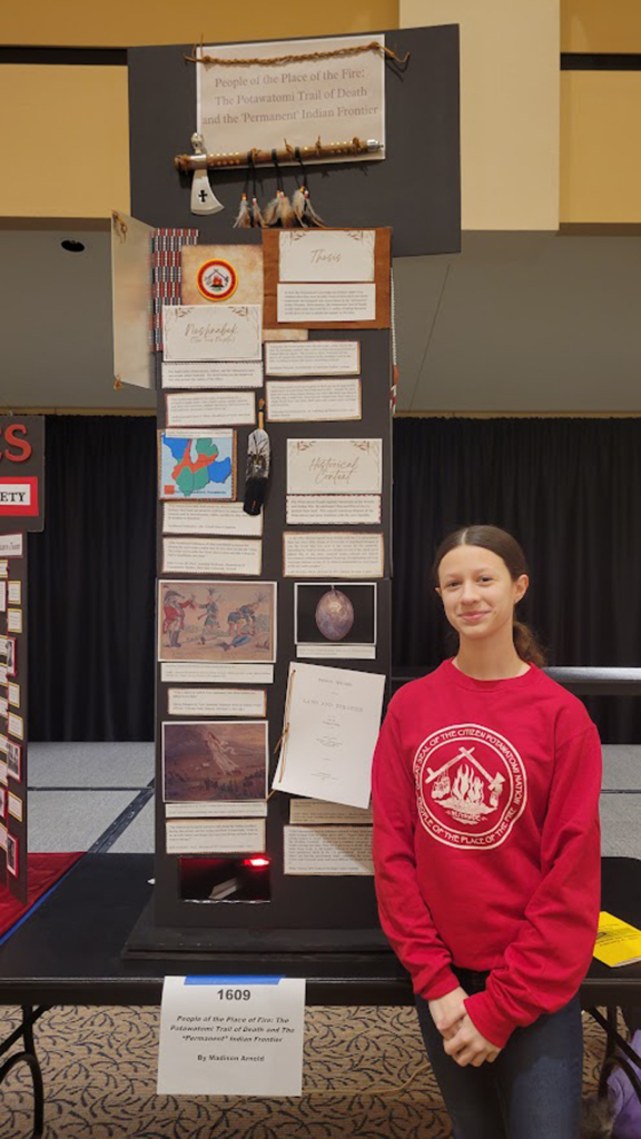Madison Arnold poses by her poster telling of the Potawatomi Trail of Death at the Kansas State History Day Competition. Madison wears a red sweatshirt with the CPN seal on it.