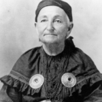 A black and white portrait of Mary Anderson Bourbonnais. She wears a ribbon shirt and layers of beaded necklaces.