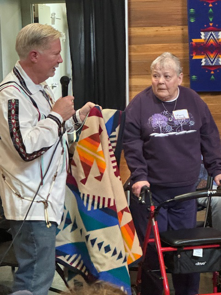 Dave Carney, holding a microphone, extends a red, orange, tan, and blue Pendleton blanket to an Elder wearing a purple sweatshirt and standing with a red walker.