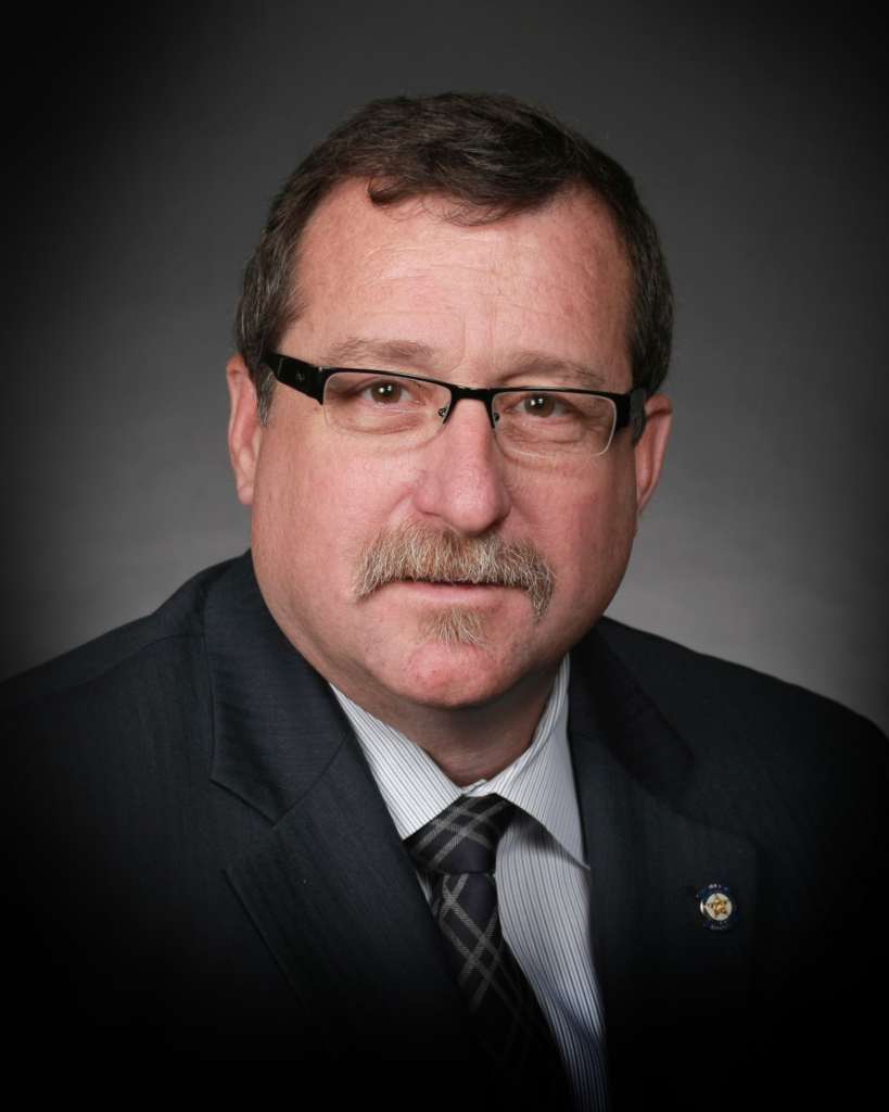 Headshot of Representative Mark McBride. He wears a suit and tie, and has dark-framed glasses on. A greying mustache matches a small goatee.