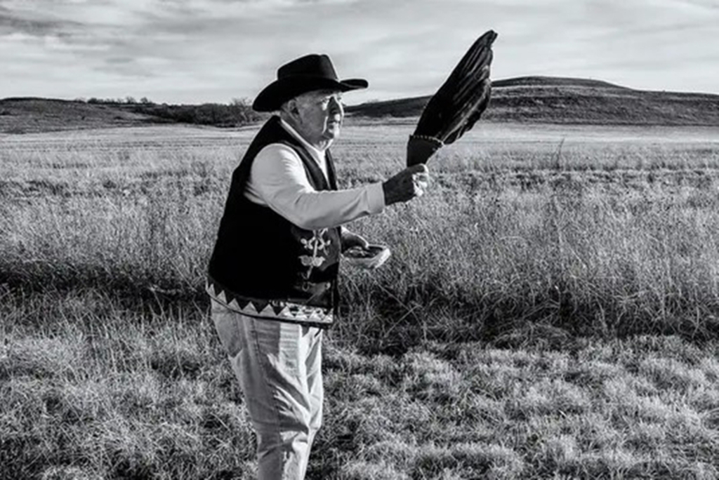 Black and white photo of Citizen Potawatomi Nation District 4 Legislator Jon Boursaw holding a fan during a blessing ceremony at Mount Mitchell. Prairie land and Mount Mitchell are visible in the background.