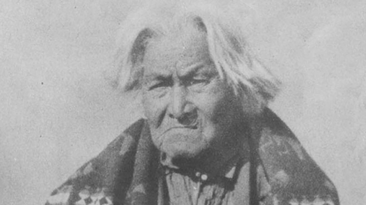 Black and white photo of Nah-Nim-Nuk-Skuk ca. 1880's-1890's. White hair falls to his ears, and he squints at the camera. A blanket is wrapped around his shoulders and his hands sit loosely in his lap.