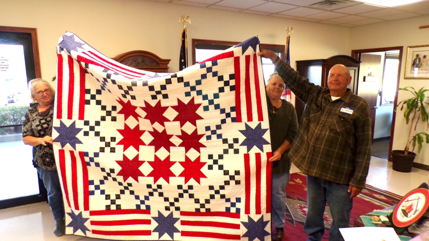 CPN Veterans and members of the Linwood, Kansas, Quilts for Veterans group hold up a large quilt with red and blue stars, red stripes, and blue and white checkers.