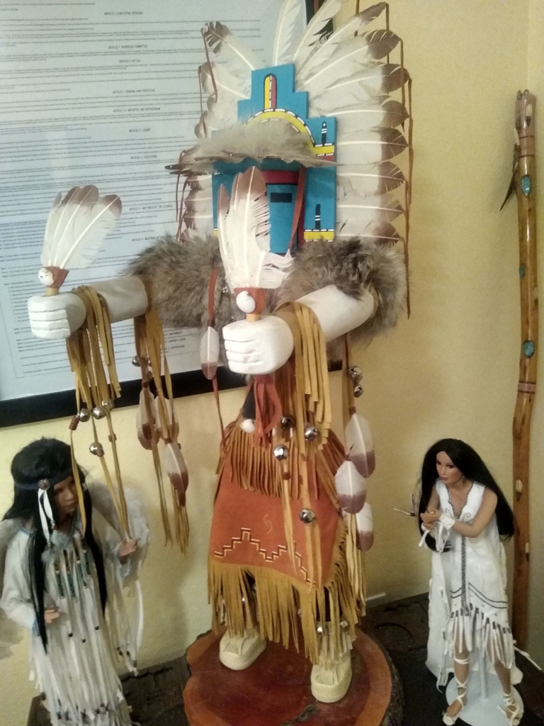 A Kachina wearing a fur shawl, feather headpiece, tassled skirt, and holding feathers in its hands.