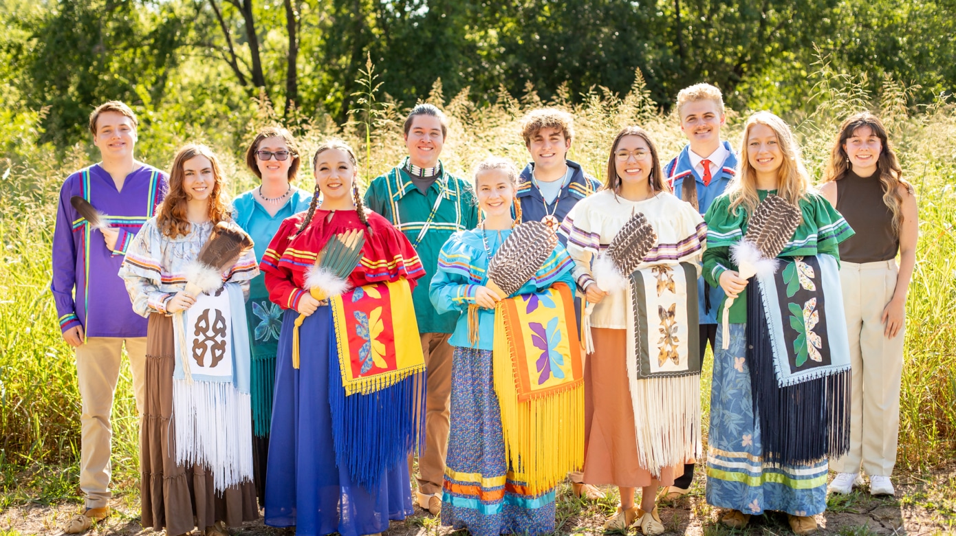 The 2022 Potawatomi Leadership class poses in afternoon sunlight. They wear colorful regalia and hold a variety of fans and eagle feathers.