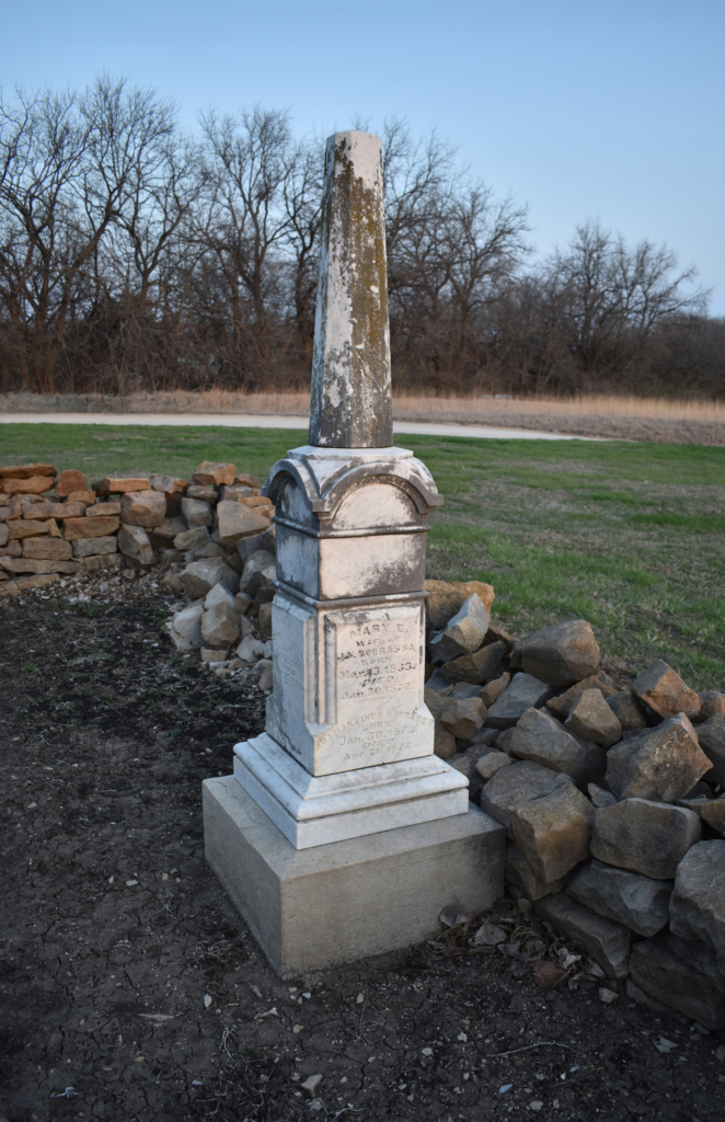 A white stone monument at the edge of a rectangular earth plot bordered by a rock wall.