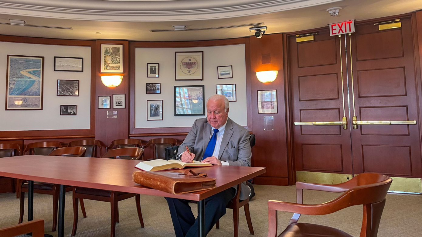 CPN Tribal Chairman John "Rocky" Barrett, wearing a grey suit and blue tie, sits at a wood desk signing a large ledger book.