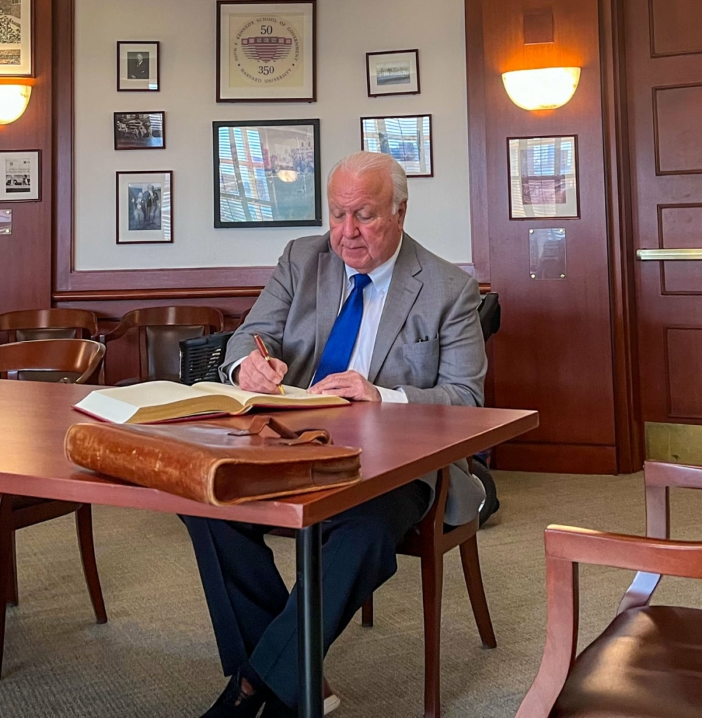 CPN Tribal Chairman John "Rocky" Barrett, wearing a grey suit and blue tie, sits at a wood desk signing a large ledger book.