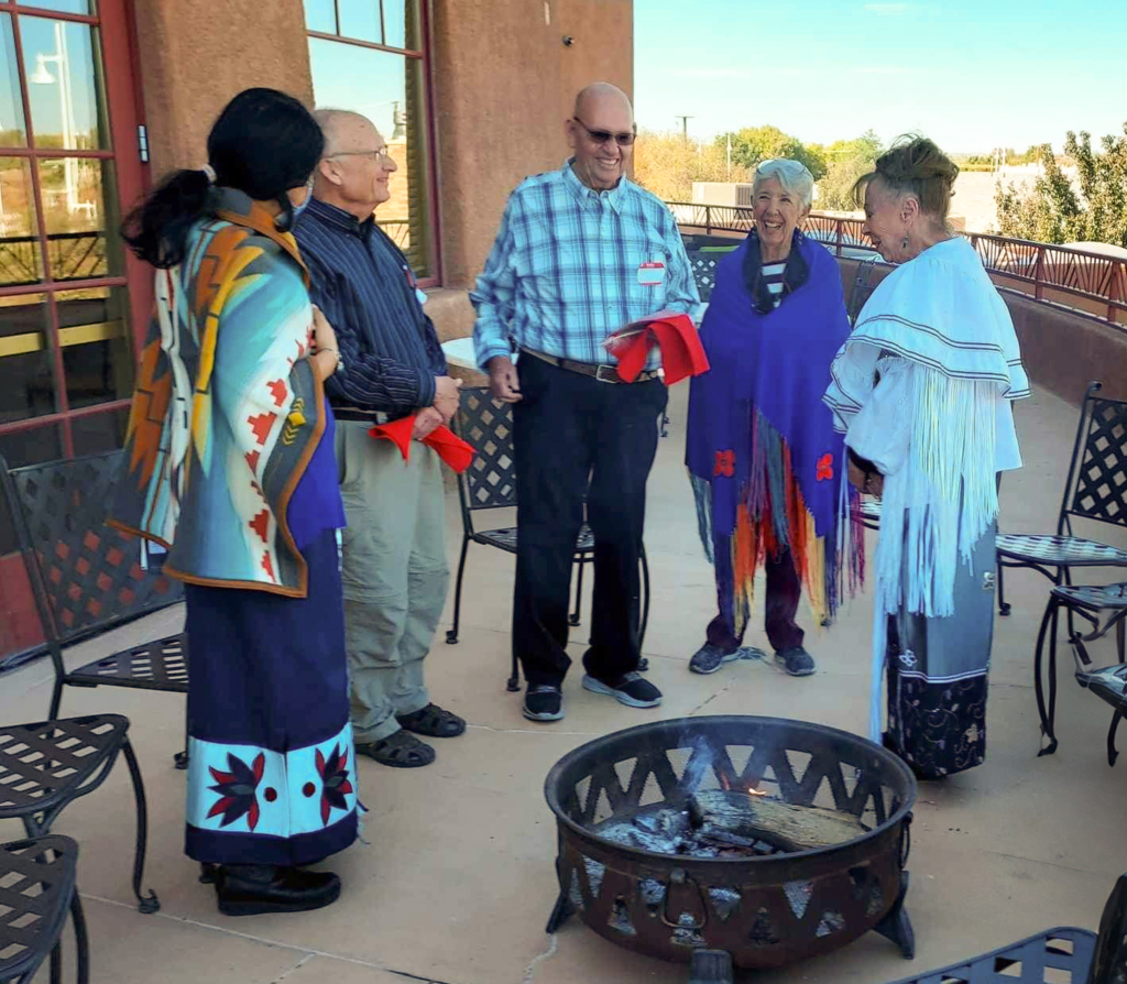 A group stands around a fire pit.Several wear shawls and ribbon skirts.