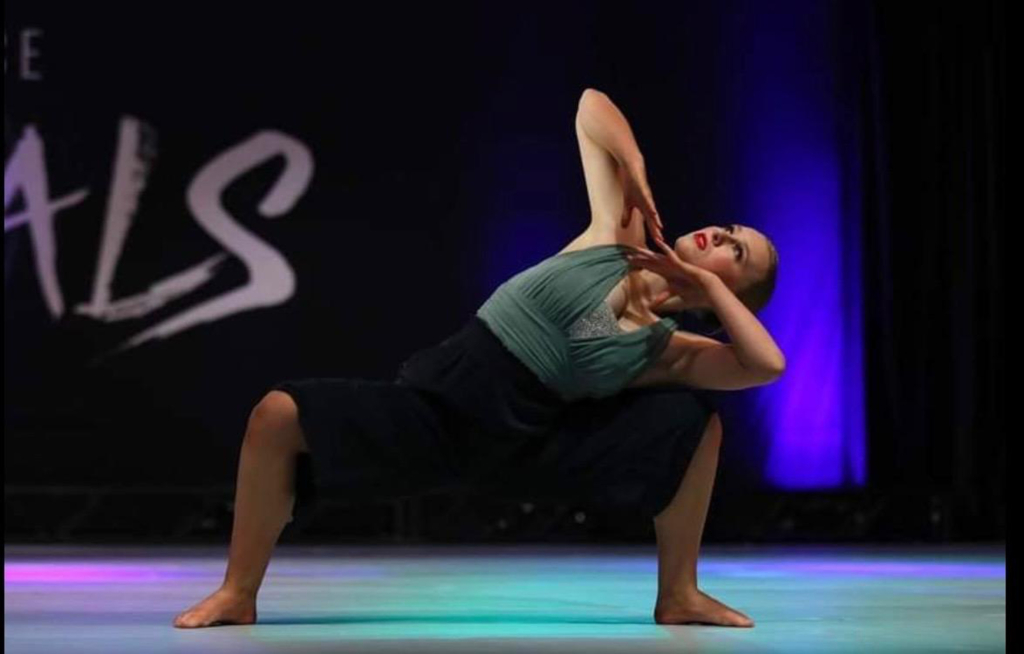 Photograph of Tribal member Piper Whitecotton dancing a solo number at on a competition stage. Standing in a deep plie in second, she leans to her left, gaze following her arms which she holds in broken T across her chest.