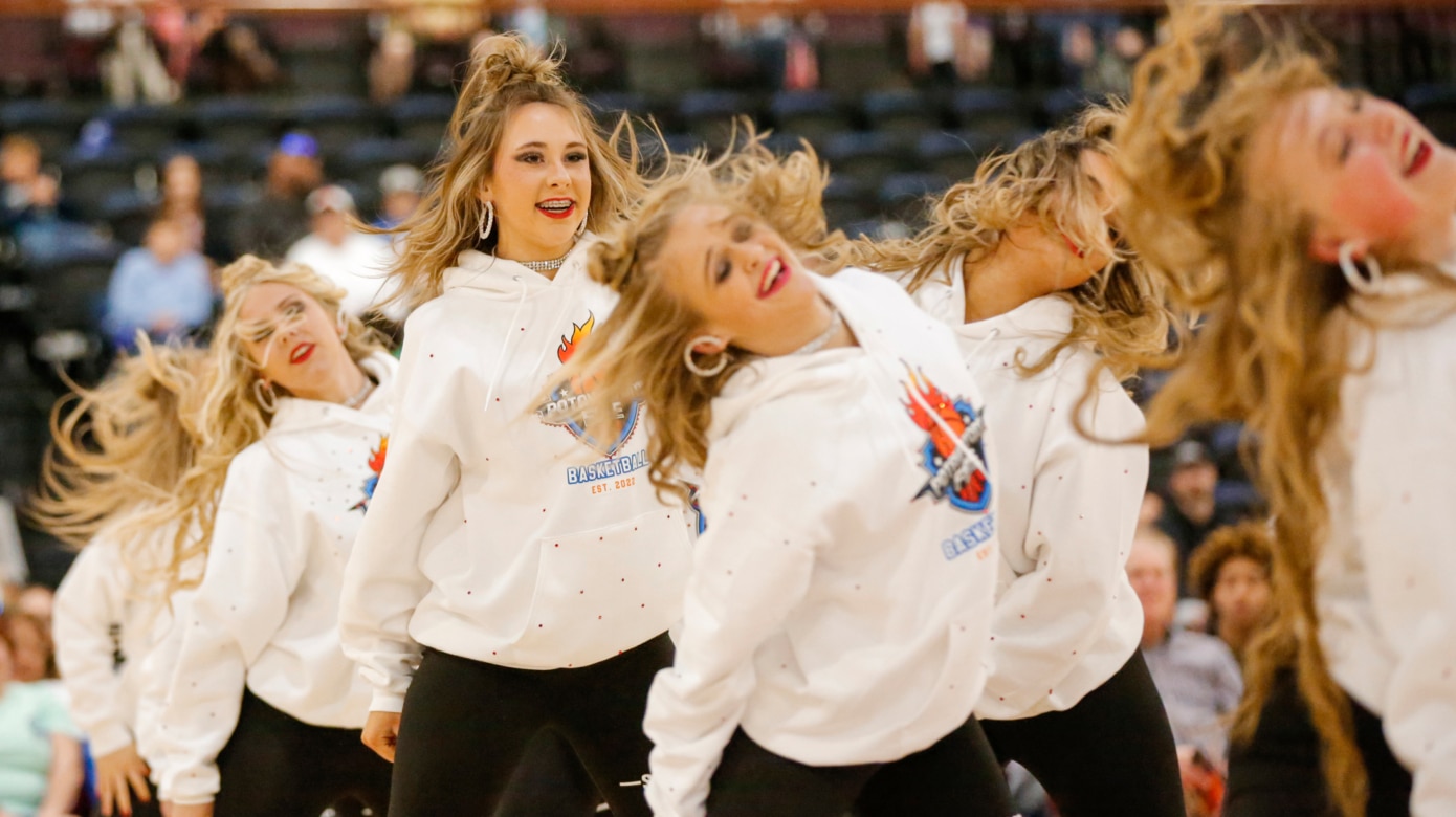 A column of dancers wearing bedazzled Potawatomi Fire hoodies perform movement in canon at their debut performance at the Potawatomi Fire's first home game at FireLake Arena on March 19, 2022. Their curled blonde hair whips across their faces, the dance movement rendered dynamic even in the still photograph.