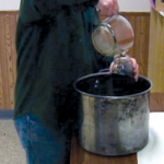 Close photo of a person in a green shirt pouring maple syrup out of a bucket into a glass jar.