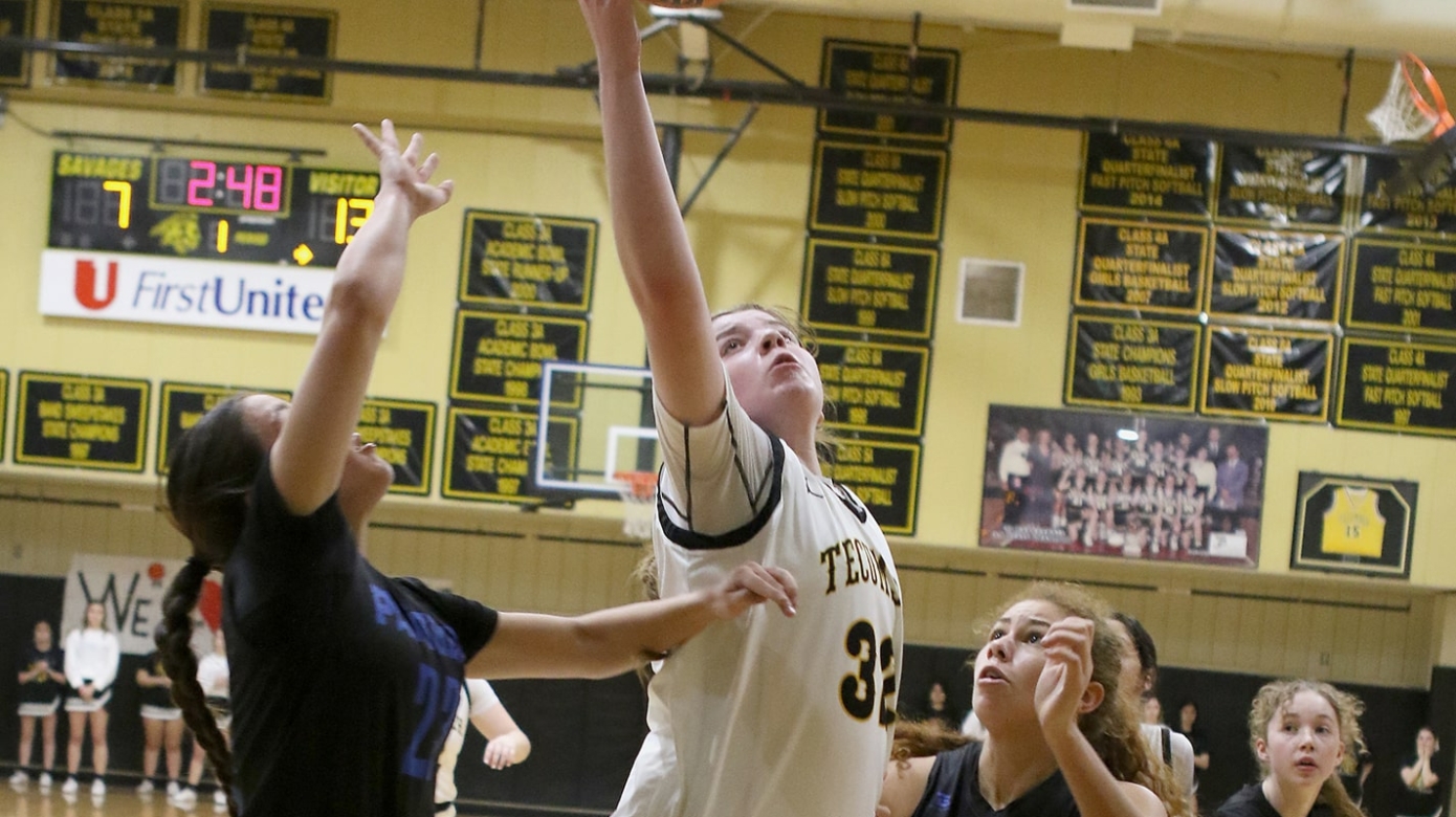A basketball player in a white uniform reaches for a rebound, surrounded by players in black uniforms.