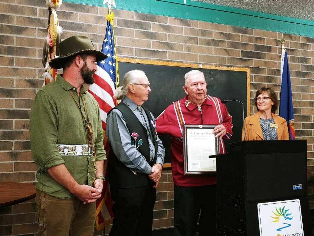 Jon Boursaw, in a red ribbon shirt, speaks at a podium. Next to him are Leg. Bob Whistler, in an applique vest, and Rep. Alan Melot, in a green shirt. They receive a framed certificate from a woman in an orange jacket.