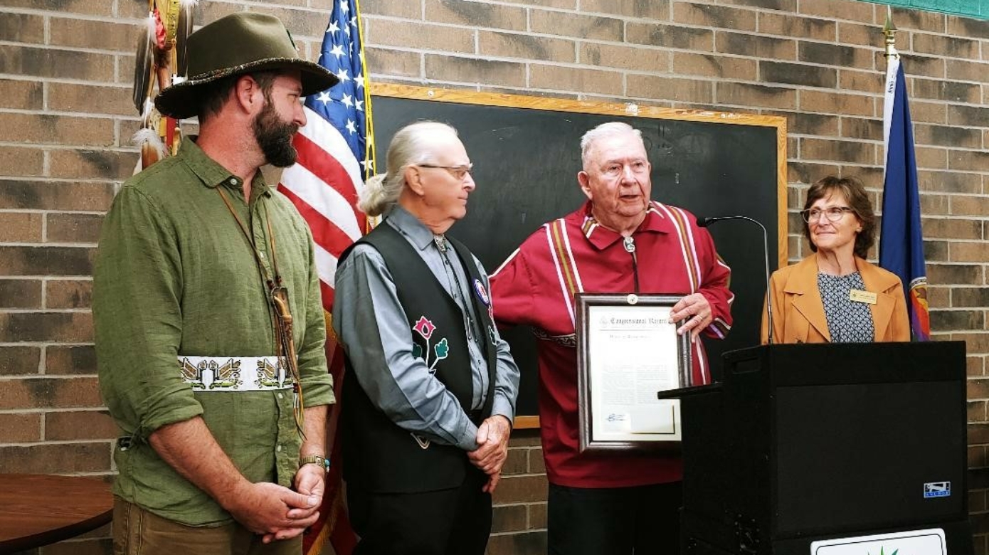 Jon Boursaw, in a red ribbon shirt, speaks at a podium. Next to him are Leg. Bob Whistler, in an applique vest, and Rep. Alan Melot, in a green shirt. They receive a framed certificate from a woman in an orange jacket.
