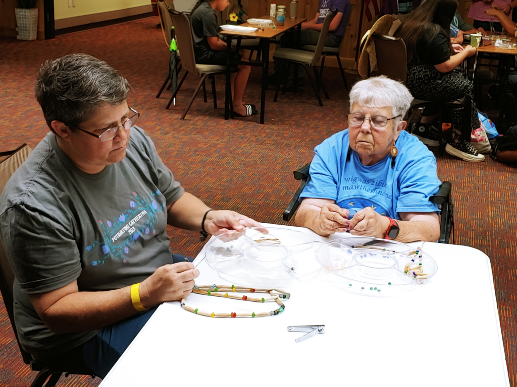 Two people with short grey and white hair sit at a table making hairpipe necklaces.