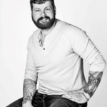 A black and white portrait of CPN artist Riley Wolery. He sits with one hand at his hip on the edge of a stool against an all white background. Tattoos cover his forearms, visible where his long shirt sleeves have been pulled up to the elbow.