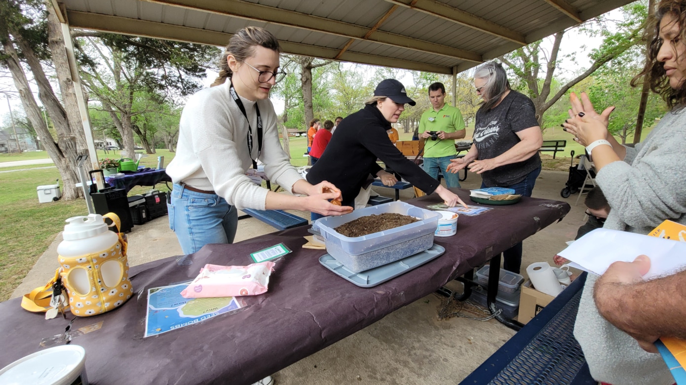 A person in a white sweater and blue jeans demonstrates making a seed bomb for planting pollinator-friendly plants at a park pavilion at a recent Earth Day event.