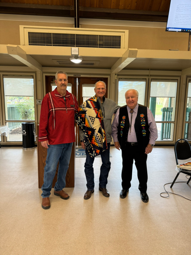Legislator Mark Johnson and Tribal Chairman John "Rocky" Barrett stand with Tribal member Marvin Stites, who is draped with a Pendleton blanket.