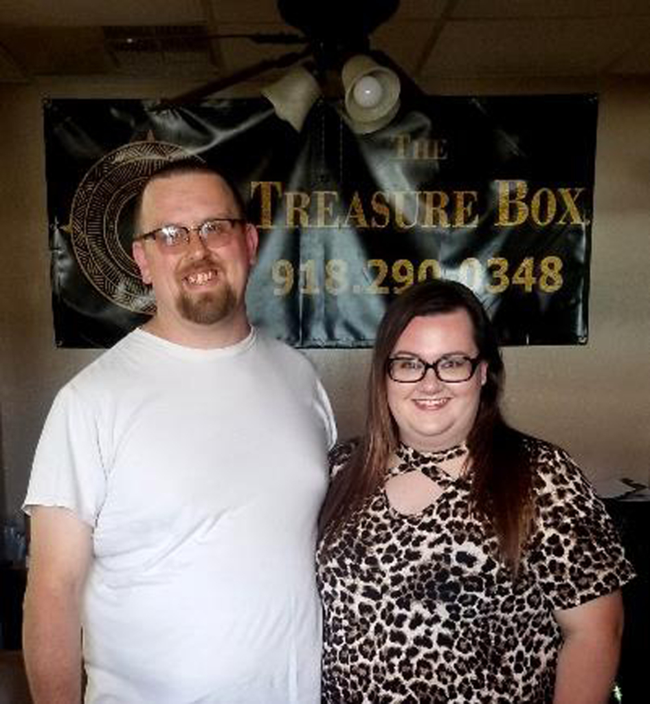 Michelle and Trent Whitson in front of a sign displaying the name of their business - The Treasure Box. 
