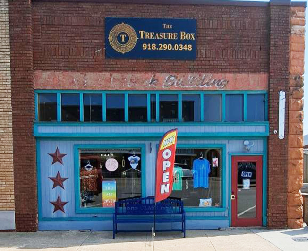 The storefront of the Treasure Box in Stroud, Oklahoma. The storefront is painted bright blue, which contrasts with the red brick of the building. A red "open" flag flies in front of the building.