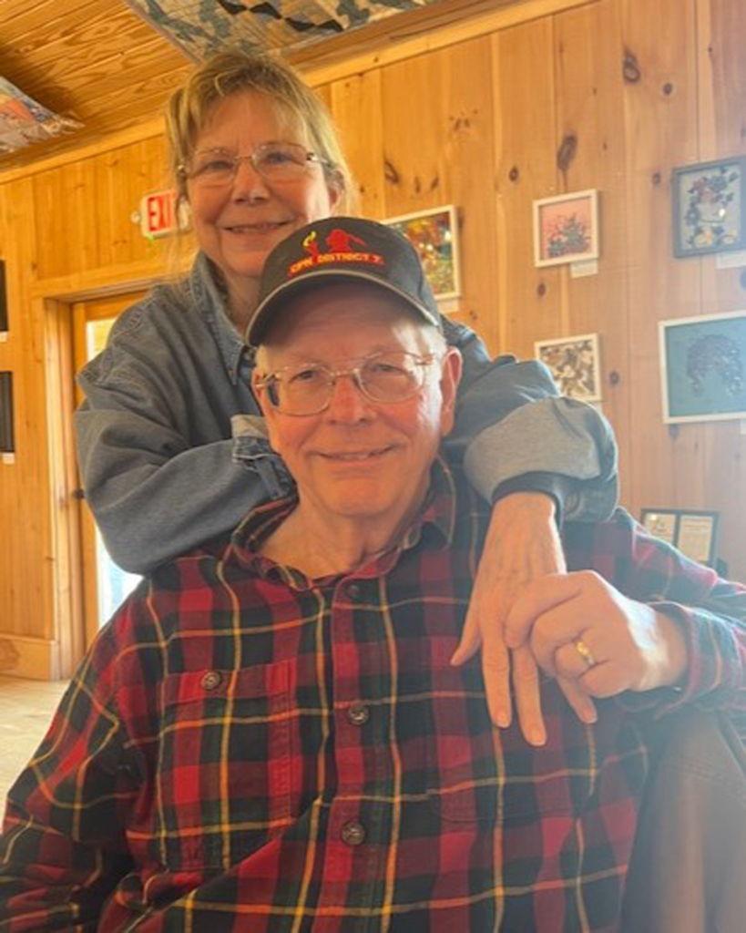 Man wearing a plaid shirt is seated in the foreground. A woman wearing a denim jacket stands behind him and wraps one arm around his shoulder.