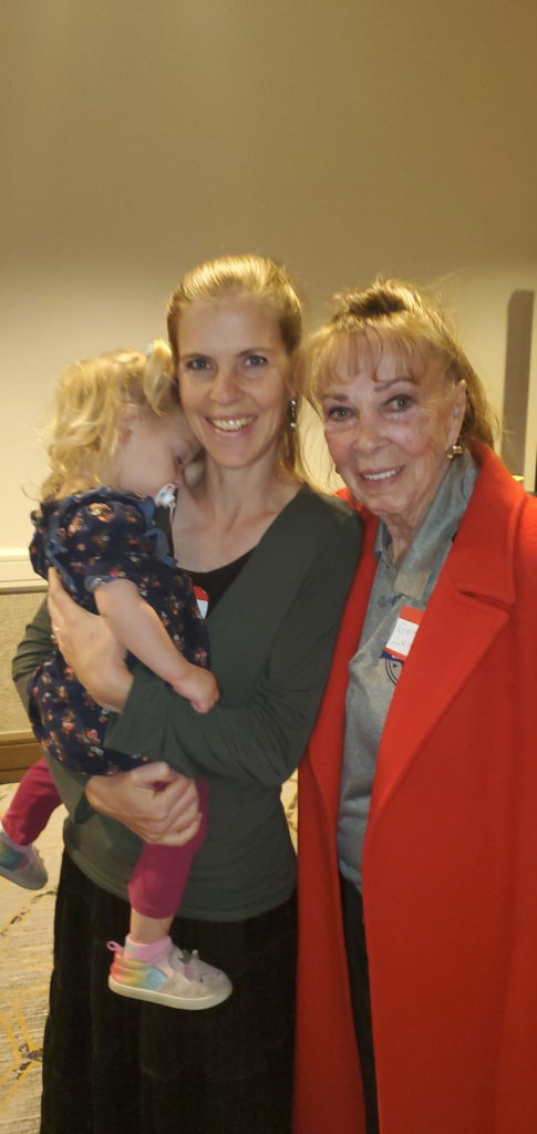 Gene Lambert, wearing a red blazer, stands next to a young mother holding a toddler with curly hair and wearing a patterned dress.