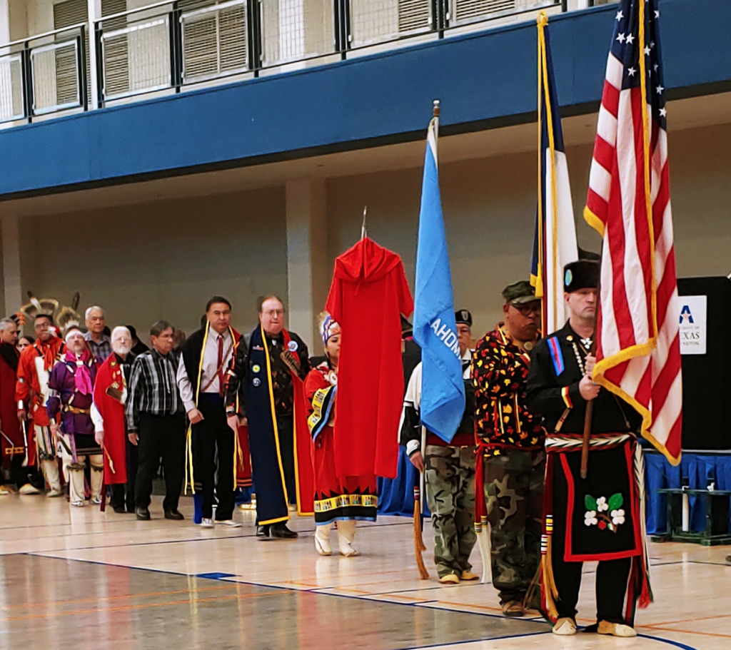 Uniformed Color Guard lead a Grand Entry procession in an indoor gymnasium. Behind the flag bearers is a person carrying a red dress to honor Missing and Murdered Indigenous Women.