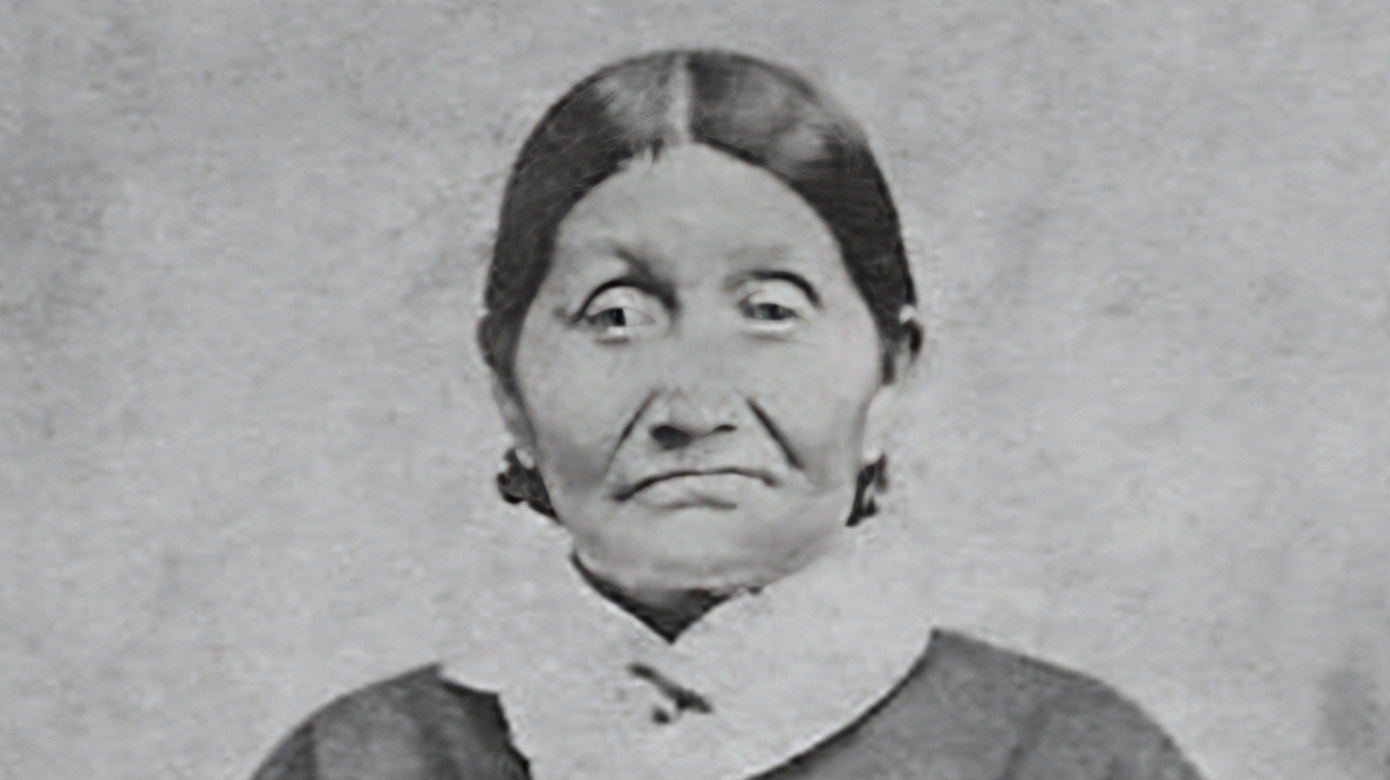 Archival portrait of Wat-che-kee, or Josette Bergeron. Hands folded at the waist, she wears a dark dress and light scarf. Her hair is parted in the middle and tied back.