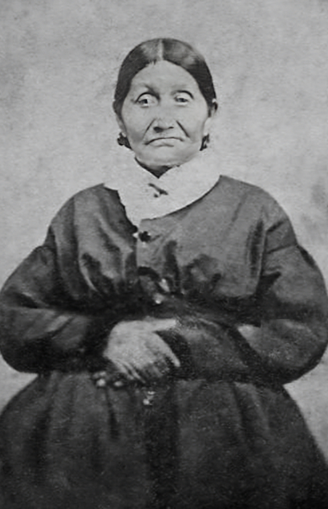 Archival portrait of Wat-che-kee, or Josette Bergeron. Hands folded at the waist, she wears a dark dress and light scarf. Her hair is parted in the middle and tied back.