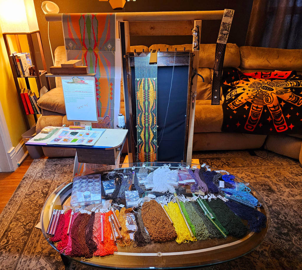 Coffee table with an array of colored beads laid out on it, before a large beading loom illuminated by a lamp and with a work in progress on it.