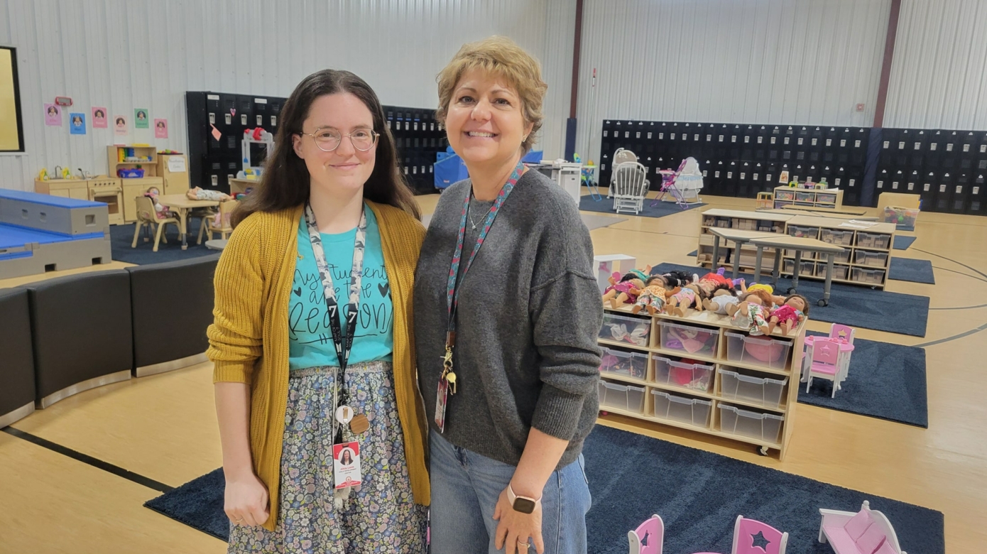 CPN After School Program teacher Alicia Lamb, wearing a blue shirt, floral skirt, and yellow cardigan stands in front of a room full of toys alongside program coordinator Jerri Mayer, wearing jeans and a grey sweater, her blonde hair short around her face.