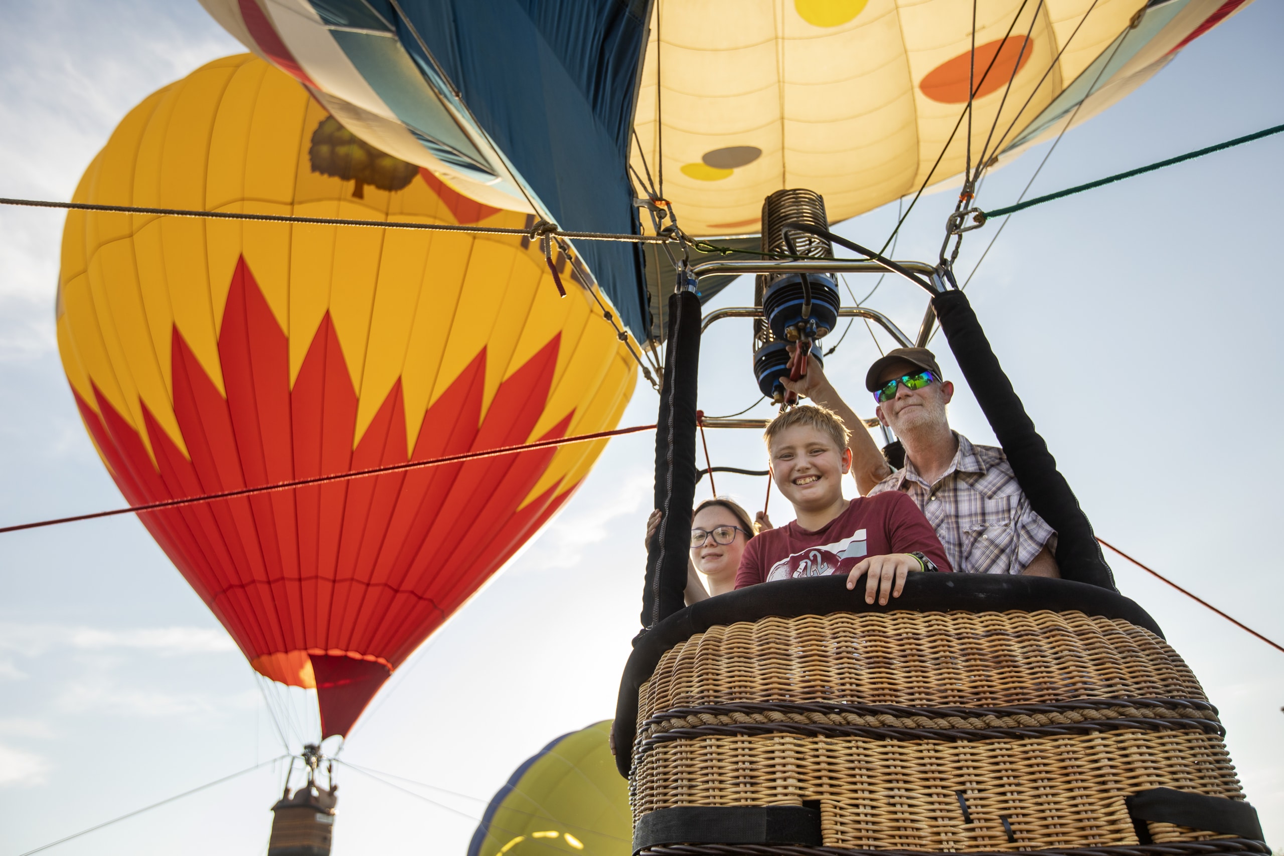 A family looks down from a hot air balloon basket, beaming. Several other hot air balloons are visible in the background.