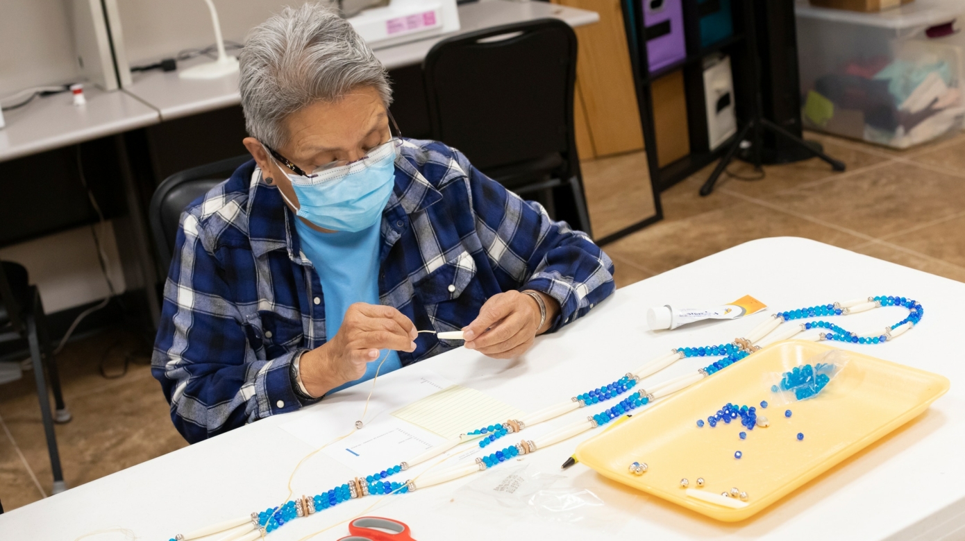 A participant in one of the CHC cultural classes carefully threads a bead in their blue and white bandoiler project under the instruction of Leslie Deer, CHC Cultural Activities Coordinator.