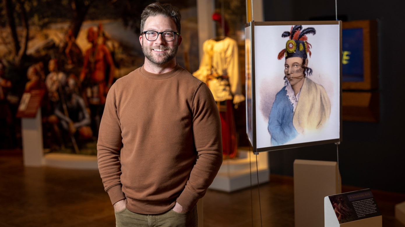Curator Blake Norton stands beside a glowing photo exhibit at the Citizen Potawatomi Nation Cultural Heritage Center. He wears a brown sweater and dark glasses.