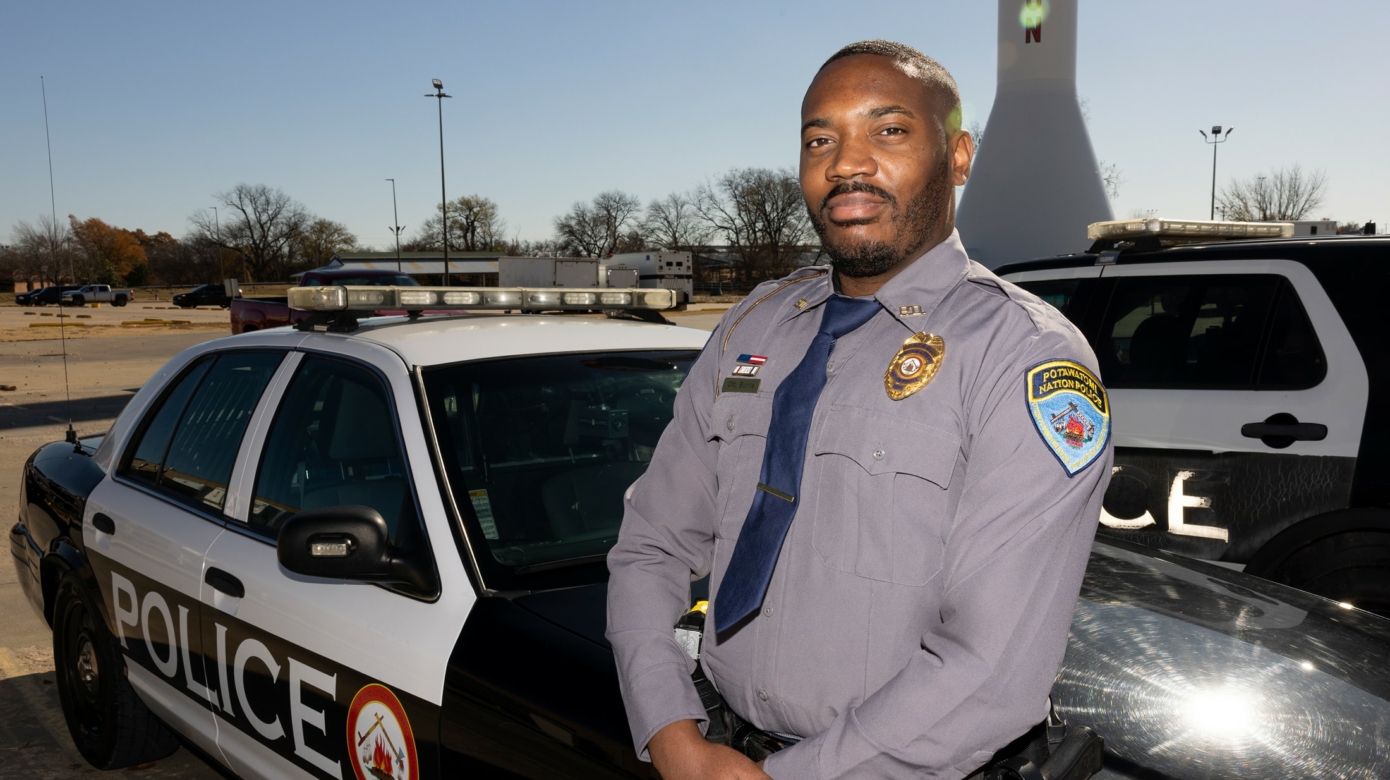 Patrol Officer Marcus Burris leans against a patrol car. He wears a uniform button down and tie, and his hands are folded at his waist.