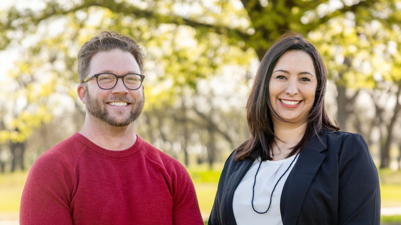 Blake Norton, wearing a red sweater and khaki pants, and Keisha Wolf, wearing jeans, a white blouse, and a black blazer, stand outside beneath sunlit trees.