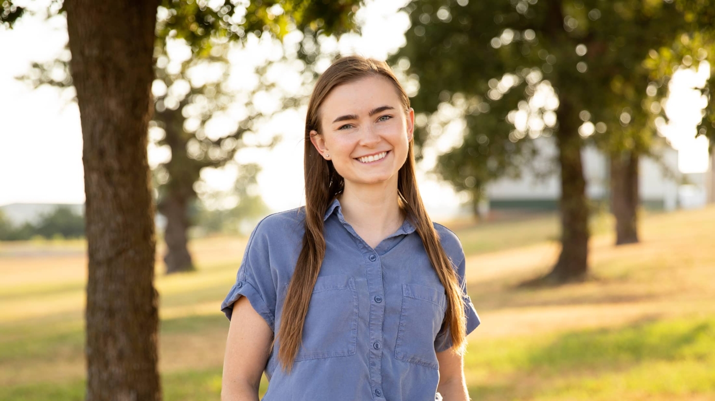 Nurse Practitioner Christine Munoz poses outside in golden afternoon light. Her light blue blouse accents her light eyes, and her brown hair falls past her shoulders. She stands with her hands in her pockets and smiling brightly at the camera.