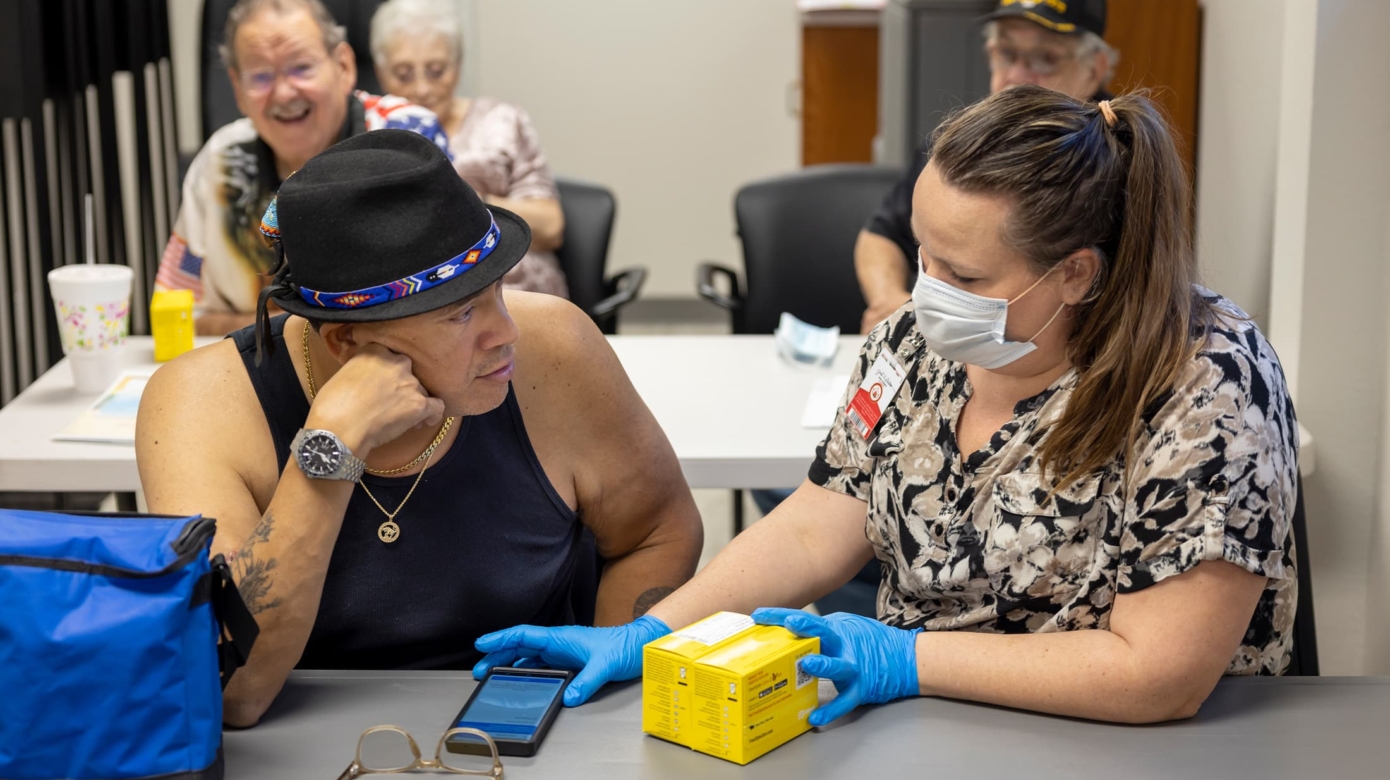 A patient in a black tank top and bead-trimmed hat sits at a table next to CPN Diabetes Coordinator Katie Brown, who demonstrates the use of glucose monitoring technologies.