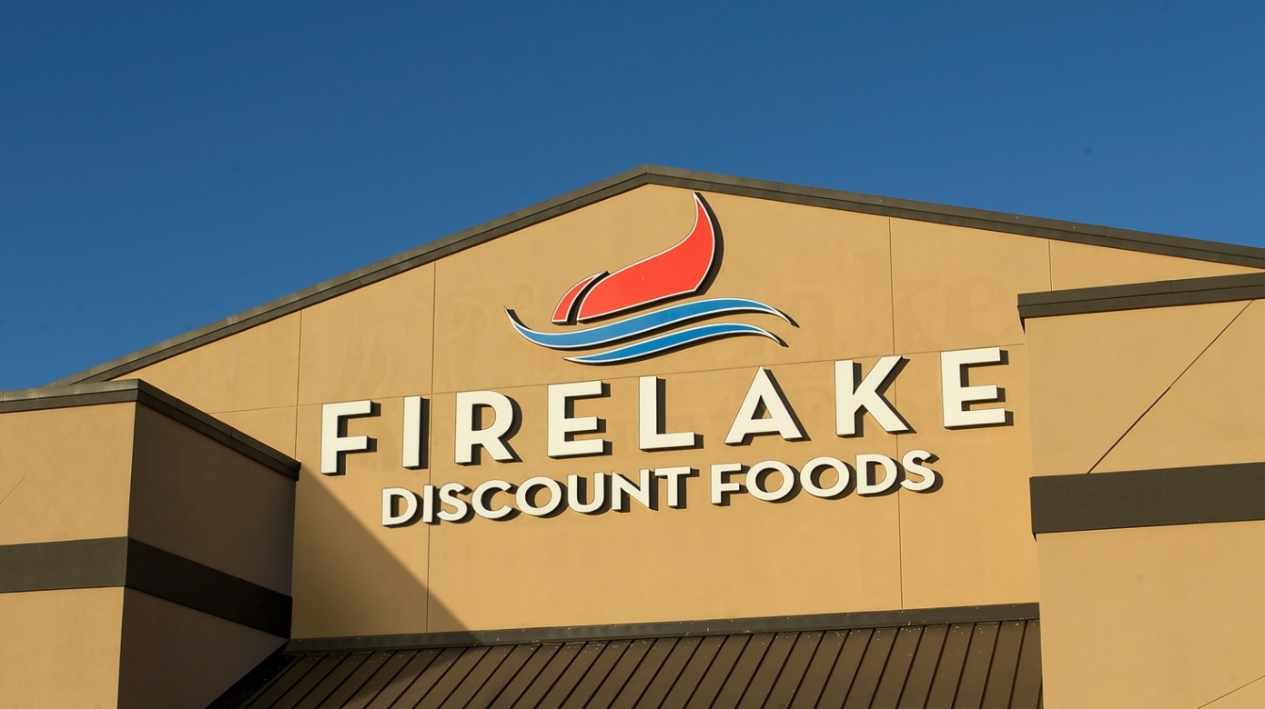 The front of FireLake Discount Foods at golden hour. A red flame rises from two blue swooped lines representing water over the name FireLake Discount Foods in white lettering on a tan building front.