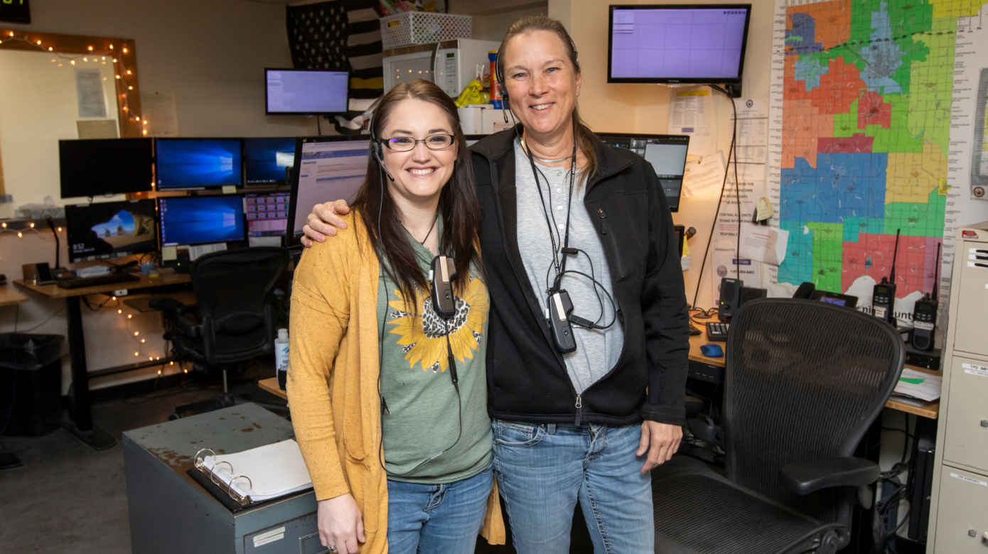Two dispatchers wearing jeans, tshirts, and cardigans pose for a photo in their command center at the CPN Tribal Police Department. Monitors and a county map are visible behind them.