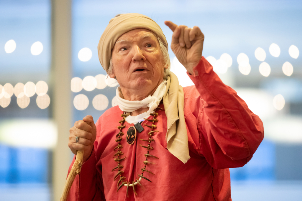 George Godfrey, wearing red handmade regalia and a beige scarf around his head, is photographed while speaking to a group.