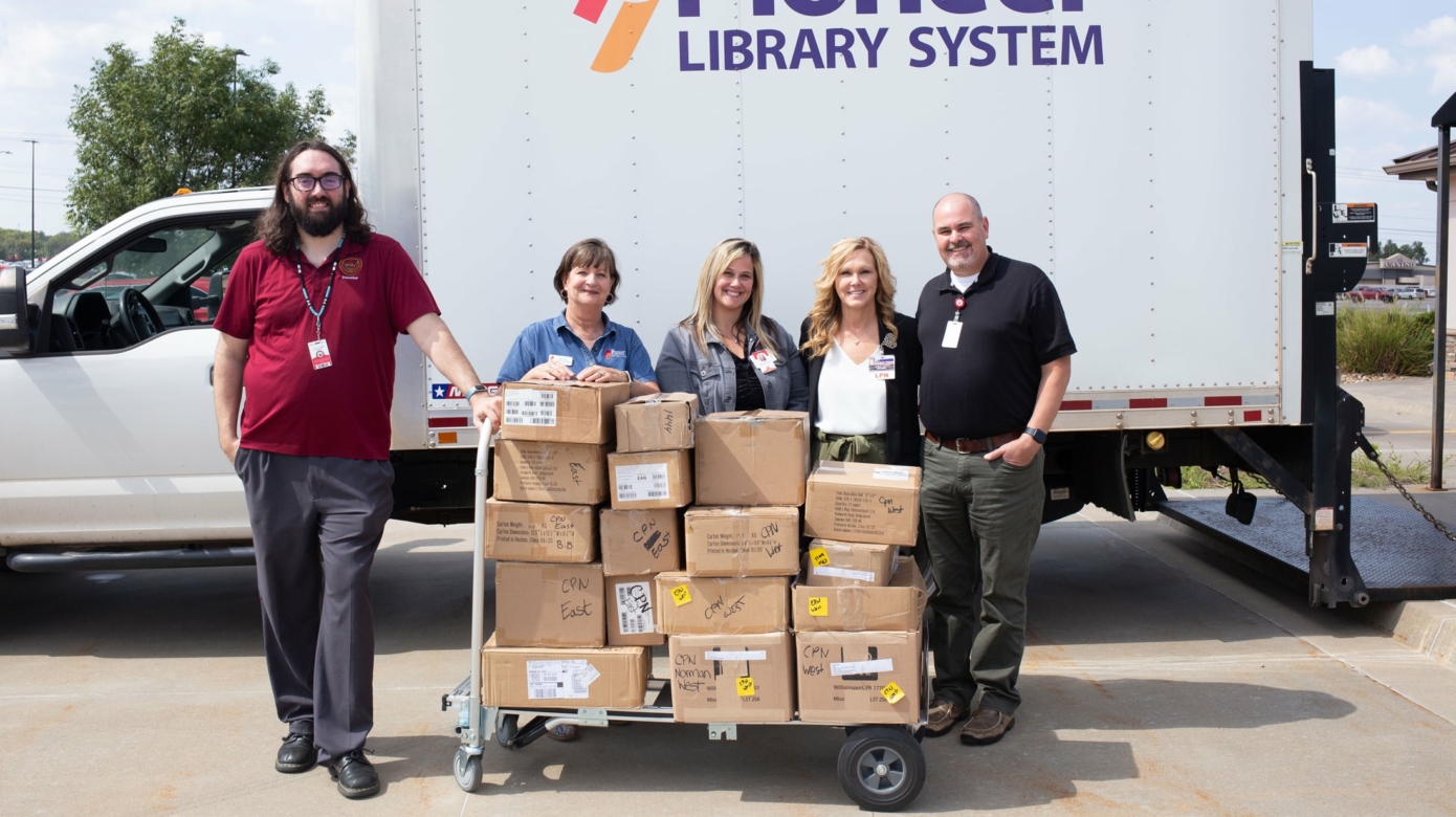 Four staff members stand with a cart loaded with cardboard boxes of books in front of a box truck labelled with the Pioneer Library System logo.