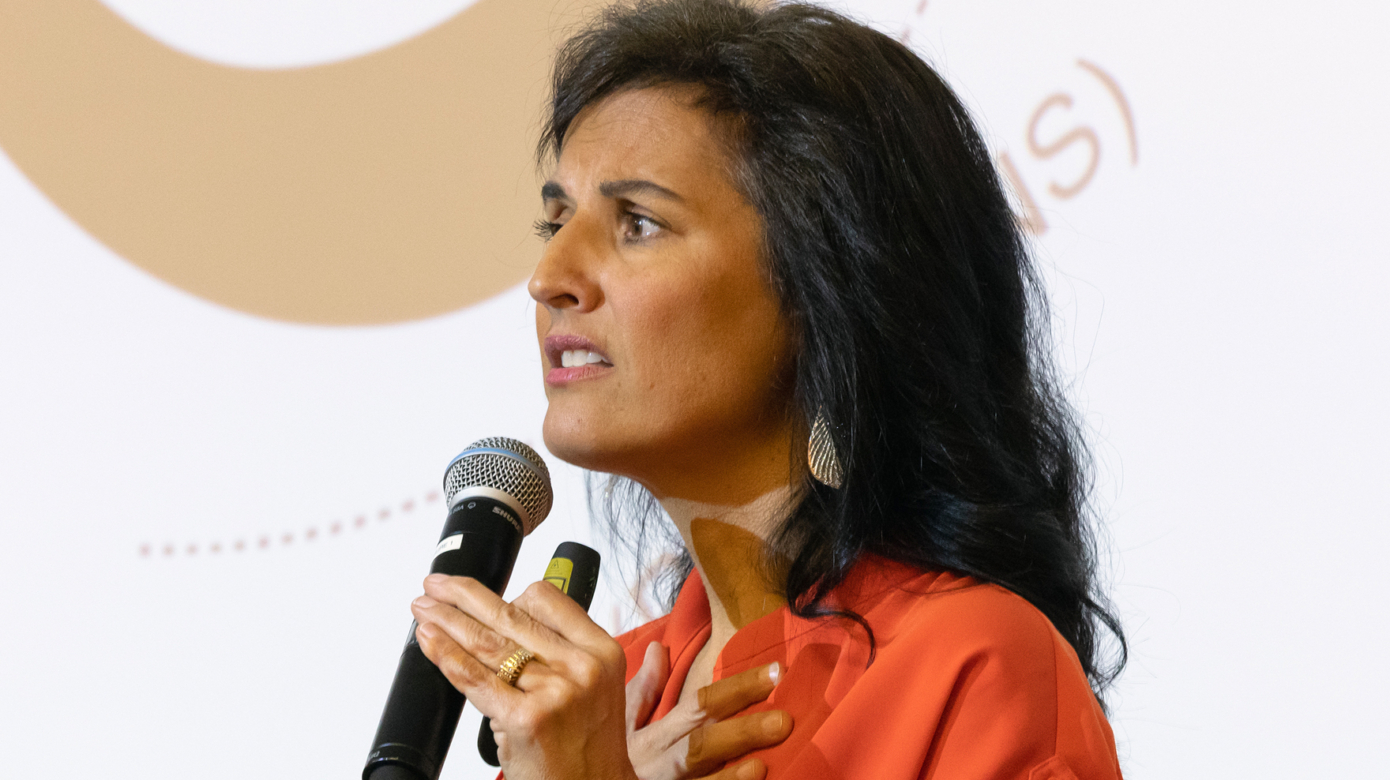 Tracy Rader stands holding a microphone in one hand, and holds her other hand over her heart. She wears a vibrant orange dress and her dark hair falls past her shoulders.