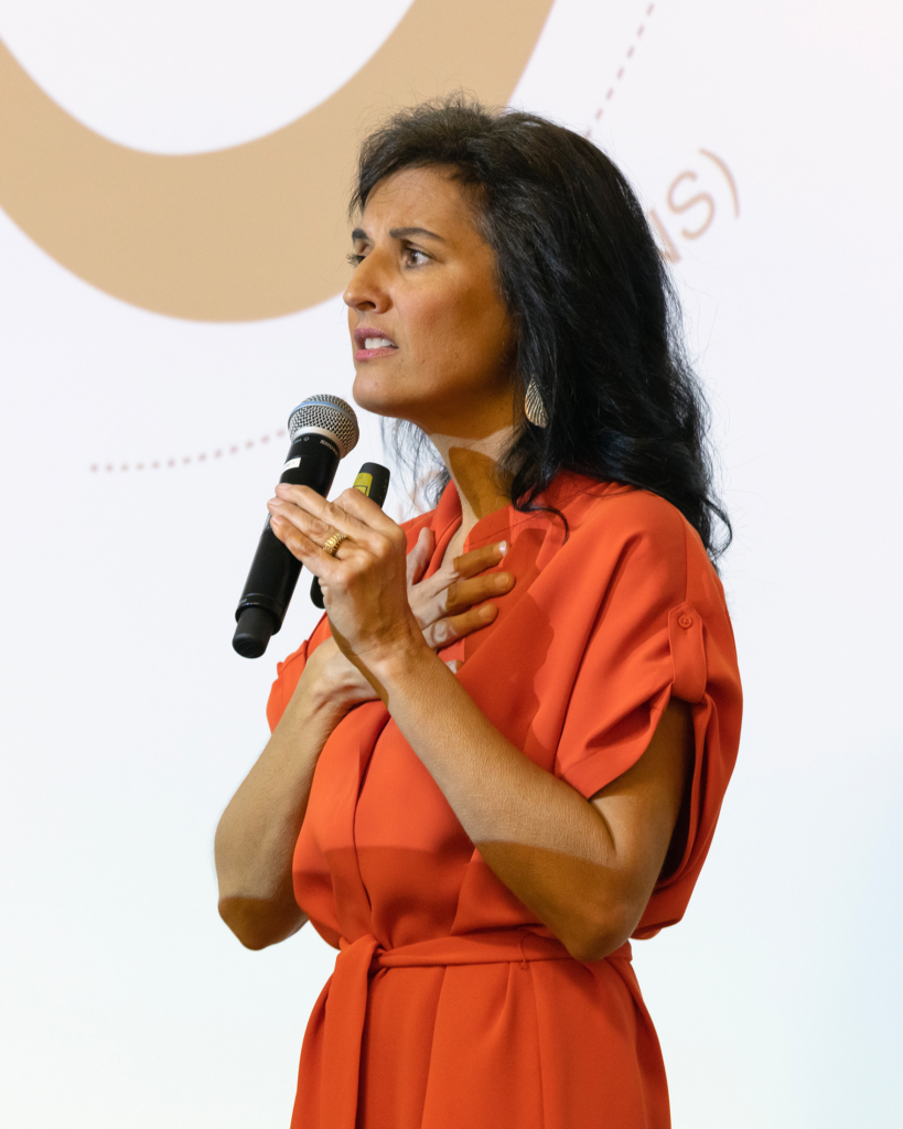 Tracy Rader stands holding a microphone in one hand, and holds her other hand over her heart. She wears a vibrant orange dress and her dark hair falls past her shoulders.
