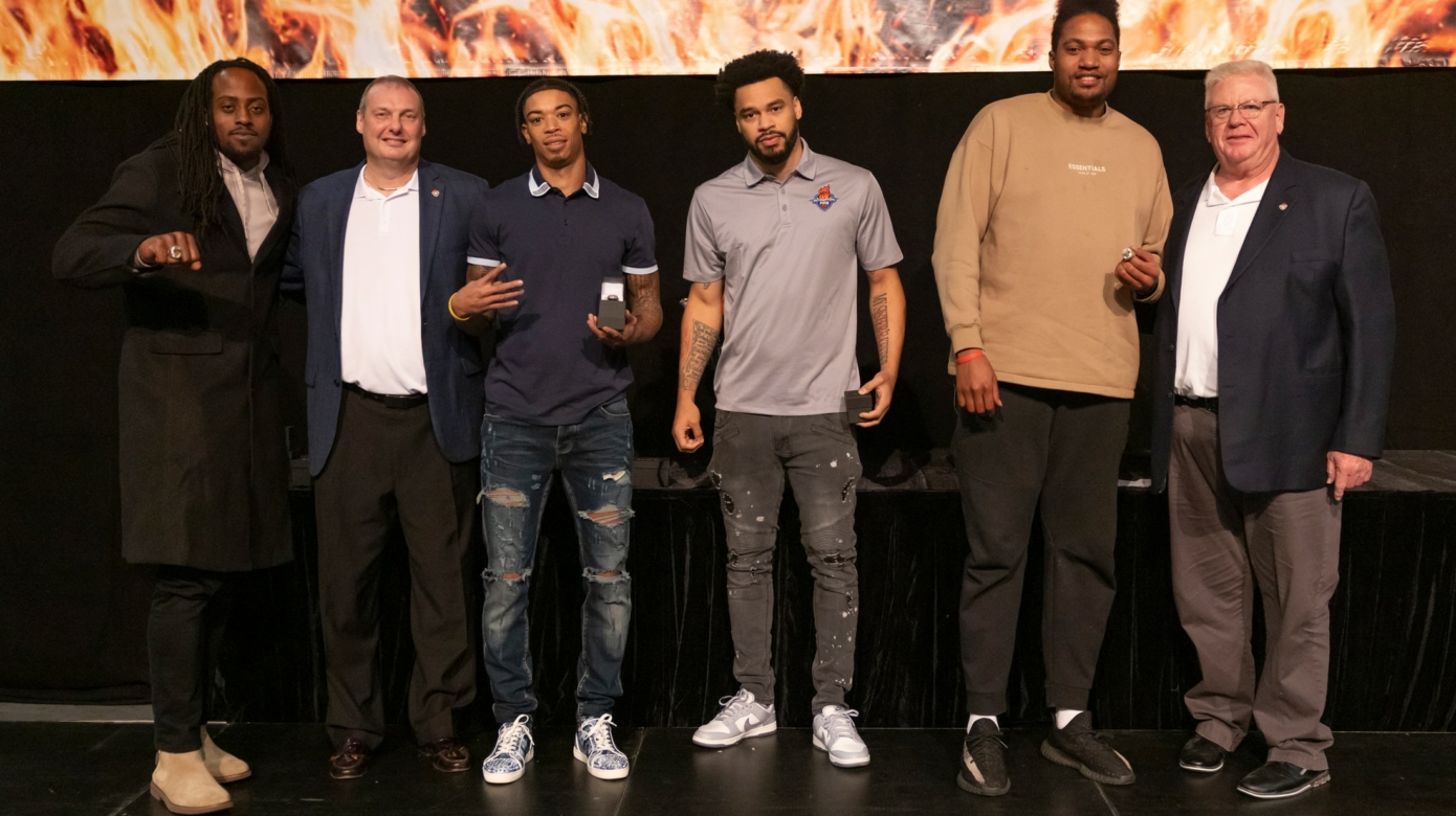 Players stand on stage with the team coach and general manager displaying their championship rings.