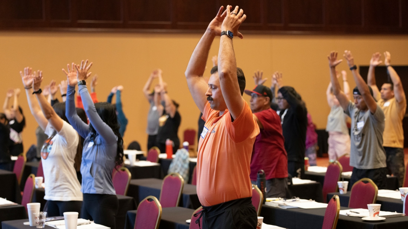 A participant with a dark mustache and wearing an orange polo and black pants raises their hands above their head in an exercise at the Native American Fitness Council conference at the Grand Casino Hotel & Resort.